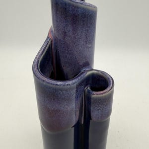 May include: A handcrafted ceramic vase in shades of deep blue and purple. The vase features a unique, curved design with multiple compartments and openings. The glossy finish enhances the rich color palette, making it a decorative accent.