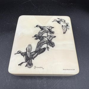 May include: A rectangular, off-white marble piece with a black and white illustration of ducks in flight. The artwork is signed "BORGEREEN '91" and includes the text "Montana Marble Inc. © 1985".