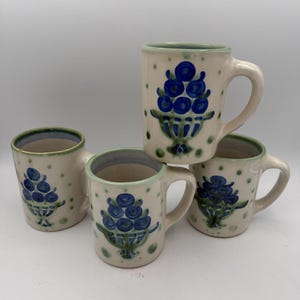 May include: A set of four off-white ceramic mugs featuring a hand-painted blue berry motif arranged in a basket shape. Each mug is decorated with small green polka dots and has a light green rim. The mugs have a cylindrical body and a side handle.