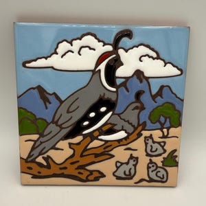 May include: Square ceramic tile featuring a stylized illustration of quail birds in a desert landscape. The birds are gray with black and white markings, perched on a brown branch. The background includes blue mountains, clouds, and a light blue sky.