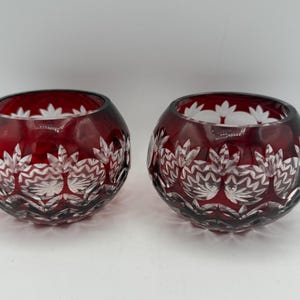 May include: Two round, deep red glass bowls featuring intricate cut-glass patterns. The exterior surface displays a repeating design of clear, faceted floral motifs and horizontal wavy lines, contrasting against the dark red glass body.