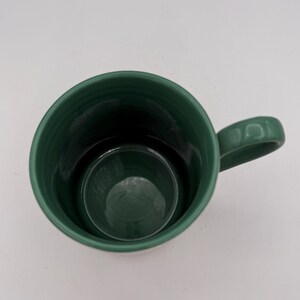 May include: A dark green ceramic mug with a handle. The mug has a cylindrical shape with a slightly flared rim. The interior of the mug is a darker shade of green. The mug is empty and sits on a white surface.