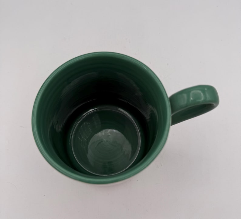 May include: A dark green ceramic mug with a handle. The mug has a cylindrical shape with a slightly flared rim. The interior of the mug is a darker shade of green. The mug is empty and sits on a white surface.