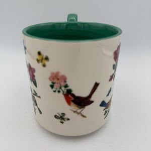 May include: Cream-colored ceramic mug with a green interior and handle. The mug is decorated with illustrations of birds, flowers, and butterflies in shades of red, pink, blue, and green. The mug is a standard size for coffee or tea.