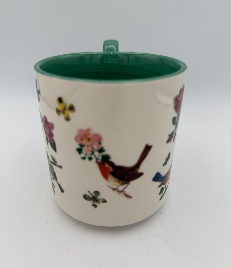 May include: Cream-colored ceramic mug with a green interior and handle. The mug is decorated with illustrations of birds, flowers, and butterflies in shades of red, pink, blue, and green. The mug is a standard size for coffee or tea.