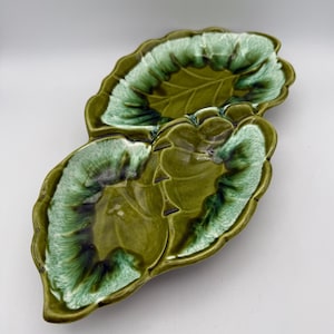 May include: A ceramic leaf-shaped serving dish with a glossy finish. The dish is a deep green color with lighter green and white accents around the edges, creating a textured appearance. The dish is designed to resemble a leaf.