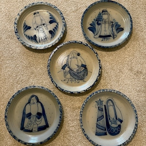 May include: A set of five round ceramic plates with a speckled blue rim and a beige center. Each plate features a hand-painted blue illustration of a bearded figure and the years 1992, 1994, 1996, 1998, and 2000 respectively, depicted in a folk art style.