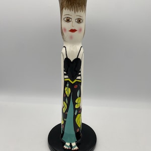 May include: A tall, decorative ceramic vase with a face design. The vase features a black top, a face with brown hair, and a black dress with leaf and floral patterns. It stands on a black circular base.