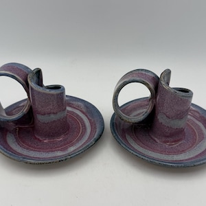 May include: Two handmade ceramic candle holders. Each has a circular base with concentric rings of purple, gray, and blue. A cylindrical candle cup rises from the center, with a curved handle. The color scheme is primarily purple and blue.