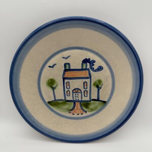 May include: A ceramic plate with a hand-painted design. The plate features a blue rim and a central illustration of a house with a brown roof, green trees, and blue accents. The plate has a diameter of approximately 8 inches.