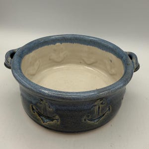 May include: A round, blue ceramic bowl with two handles. The bowl has a light interior and a dark blue exterior with embossed anchor designs. The rim is a lighter shade of blue. The bowl is suitable for serving or display.