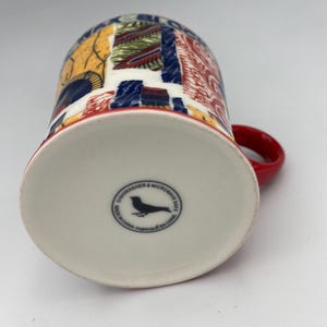 K&ouml;nnte beinhalten: Eine wei&szlig;e Keramiktasse mit rotem Henkel. Die Tasse hat ein farbenfrohes, abstraktes Design in Rot, Blau, Gelb und Gr&uuml;n. Auf der Unterseite der Tasse befindet sich ein schwarzes Vogellogo und der Text "Dishwasher & Microwave Safe. Made in China. Fabriqu&eacute; en Chine."