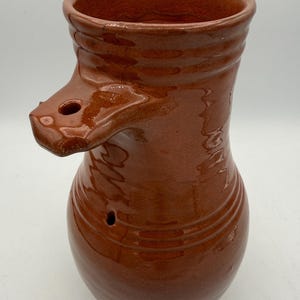 May include: A handmade, brown ceramic pitcher with a unique spout and handle design. The pitcher has a rounded base and a wide opening at the top. The exterior has a glossy finish and visible ridges.