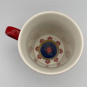 K&ouml;nnte beinhalten: Rote und wei&szlig;e Keramiktasse mit einem blauen und gelben Design im Inneren. Das Design zeigt einen Kreis mit einer roten Mitte und blauen und gelben Ringen.