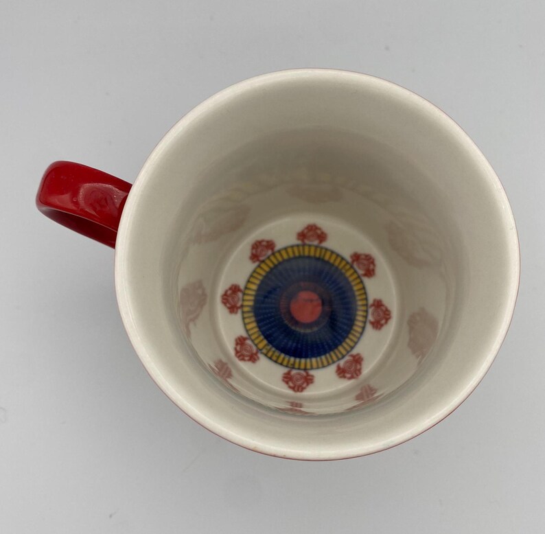 K&ouml;nnte beinhalten: Rote und wei&szlig;e Keramiktasse mit einem blauen und gelben Design im Inneren. Das Design zeigt einen Kreis mit einer roten Mitte und blauen und gelben Ringen.