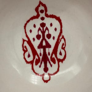 May include: Close-up of a white surface with a decorative red design. The design is symmetrical, featuring a crown-like shape with intricate details and a central vertical element. The design is likely a decorative element.