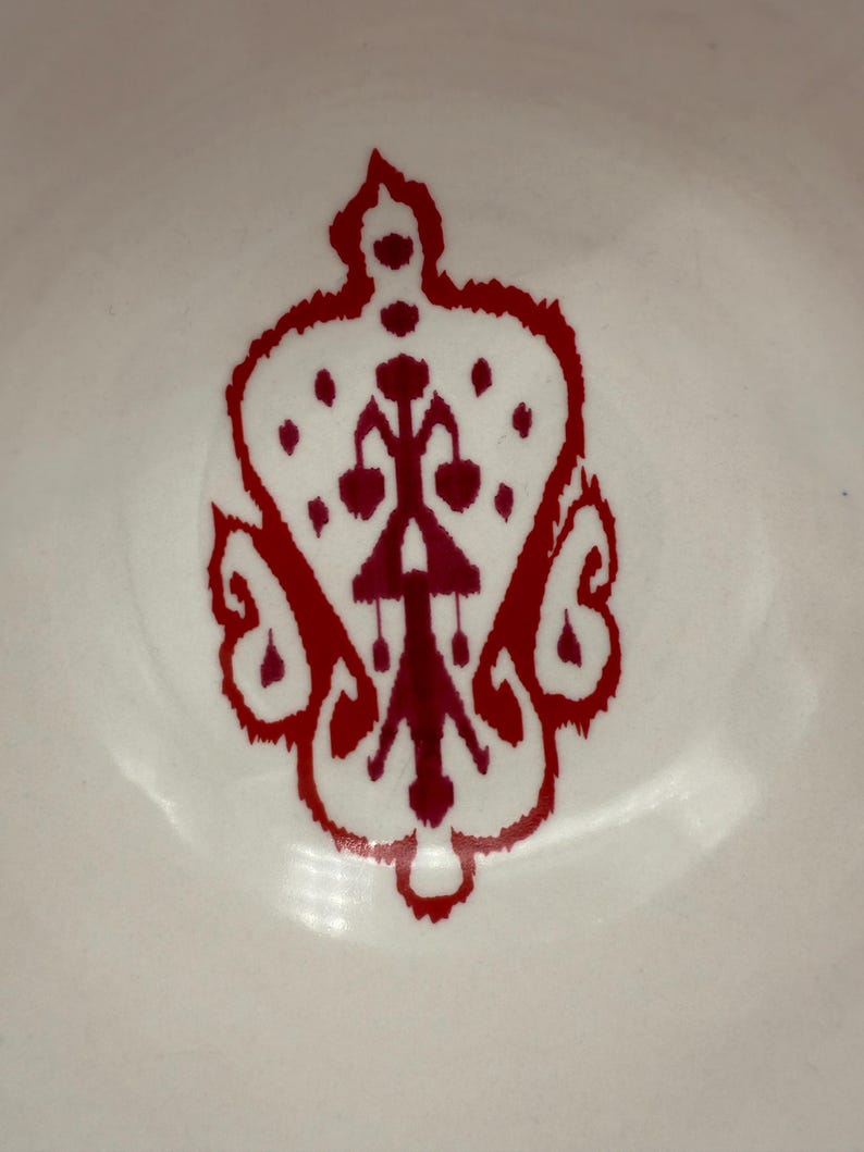 May include: Close-up of a white surface with a decorative red design. The design is symmetrical, featuring a crown-like shape with intricate details and a central vertical element. The design is likely a decorative element.