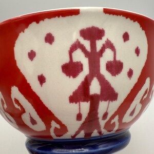 May include: A ceramic bowl with a red and white design. The bowl features a red exterior with a white interior, adorned with a red abstract pattern. The base is a solid blue color. The bowl is a decorative piece.