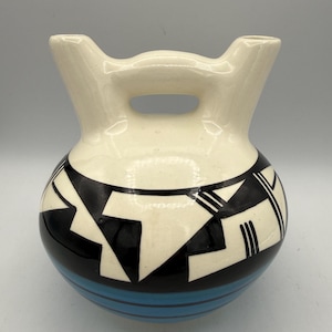 May include: A ceramic vessel featuring a rounded body with two upward-pointing spouts connected by a central handle. The upper portion is cream-colored with black geometric patterns, while the lower section displays horizontal blue stripes.