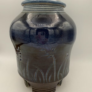 May include: A handcrafted ceramic urn with a dark blue and brown glaze. The urn features a lid and is decorated with etched grass-like designs. The base has three small feet.