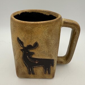 May include: A tan ceramic mug with a square shape and a large handle. The mug features a black silhouette of a moose. The interior of the mug is black.