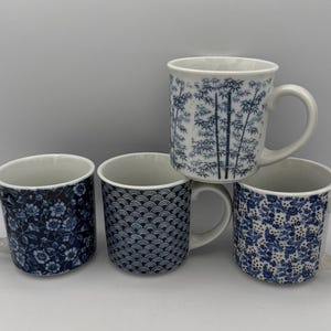 May include: Four ceramic mugs with white interiors and handles. The mugs feature blue and white patterns, including floral, bamboo, and wave designs. The mugs are arranged on a white surface.