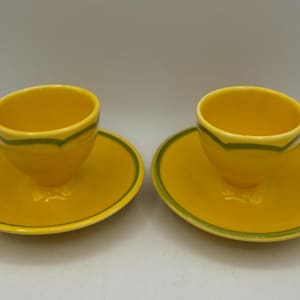 May include: Two yellow ceramic cups and saucers. Each cup has a green trim around the rim, and each saucer has a green border. The cups sit atop the saucers, creating a matching set. The set is on a white background.