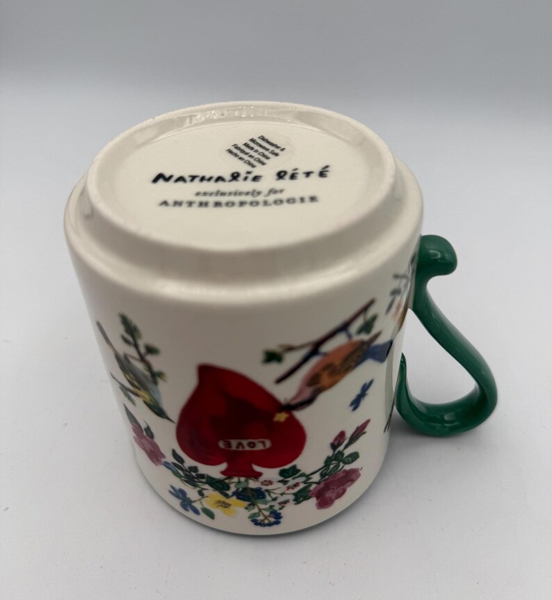 May include: White ceramic mug with a green handle, featuring a red heart with the word "LOVE" and floral and bird illustrations. The bottom of the mug has the text "Nathalie Lete" and "exclusively for Anthropologie".