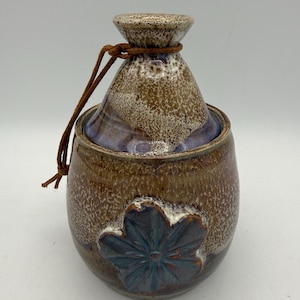 May include: A ceramic vessel with a rounded body and a tapered neck, featuring a brown and white speckled glaze. A dark blue, molded flower relief is centered on the front. A thin brown leather cord is tied around the neck of the vessel.