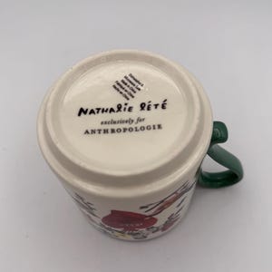 May include: A white ceramic mug with a green handle, featuring floral illustrations in red and other colors. The bottom of the mug displays the text "Nathalie Lete" and "exclusively for Anthropologie".