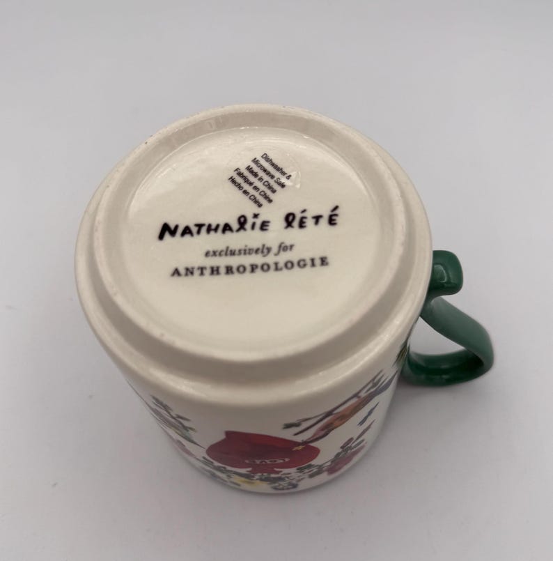 May include: A white ceramic mug with a green handle, featuring floral illustrations in red and other colors. The bottom of the mug displays the text "Nathalie Lete" and "exclusively for Anthropologie".