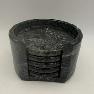 May include: A set of five circular dark gray marble coasters stacked inside a matching cylindrical marble holder. The stone features natural white veining throughout the polished surface, with a cutout section on the holder for easy access.