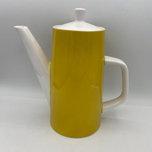 May include: A yellow and white ceramic coffee pot with a lid and handle. The pot has a cylindrical body with a white spout and handle. The lid is white with a small knob. The coffee pot is designed for brewing and serving coffee.