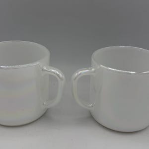 May include: Two white ceramic mugs with rounded bodies and attached handles. The rims of the mugs have a subtle iridescent sheen. The mugs are displayed against a plain white background.
