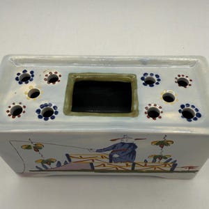 May include: Rectangular ceramic flower frog with a light blue glaze. The top has multiple holes with dark blue and brown accents. The front features a painted scene of a person fishing. The center has a rectangular opening.