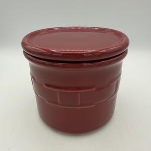 May include: A maroon ceramic jar with a matching lid. The jar has a cylindrical shape with a textured, brick-like pattern around the middle. The lid sits snugly on top, creating a cohesive and functional design. This jar is suitable for kitchen storage.