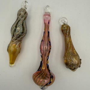 May include: Three hand-blown glass ornaments with hanging loops. The left ornament features swirling brown and amber tones. The center piece is elongated with pink, blue, and gold stripes. The right ornament has a textured brown and gold pattern.