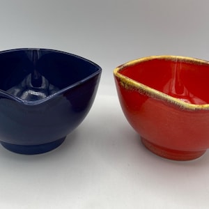 May include: Two ceramic bowls, one blue and one red. The bowls are square shaped with rounded edges and a slight lip.