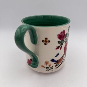 May include: Cream-colored ceramic mug with a green handle and rim. The mug is decorated with floral and bird illustrations in red, blue, and orange. A small, brown and yellow flower is also featured on the mug.