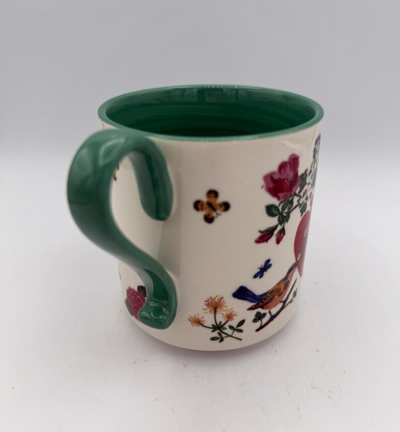 May include: Cream-colored ceramic mug with a green handle and rim. The mug is decorated with floral and bird illustrations in red, blue, and orange. A small, brown and yellow flower is also featured on the mug.