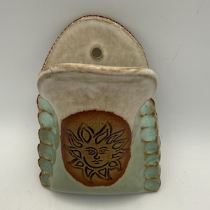 May include: A wall-mounted ceramic pocket planter featuring a cream-colored upper section with a hanging hole and a central brown circular medallion embossed with a sun face design. The sides are textured with light blue, scalloped ceramic accents.