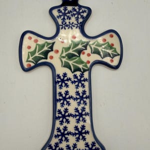 May include: A ceramic cross-shaped ornament with a blue border. The ornament features a cream-colored background with green holly leaves, red berries, and blue snowflakes. A hole is at the top for hanging.