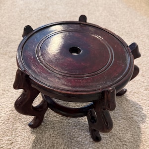 May include: A dark brown, circular wooden display stand featuring a smooth, polished top surface with a central circular cutout. The stand is supported by five curved, ornate legs that extend outward, providing a stable base for the elevated platform.