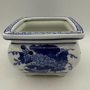May include: A square ceramic planter with a white base and blue hand-painted grape and vine motifs. The rim features a decorative blue scroll pattern, and the vessel sits on four small feet, showcasing a glossy finish and a wide, open top.