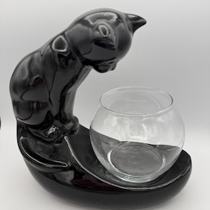 May include: A glossy black ceramic cat figurine with a curved body, positioned to look down into a clear, round glass bowl that is integrated into the base. The cat has smooth, reflective surfaces and a rounded head, while the glass bowl is transparent.