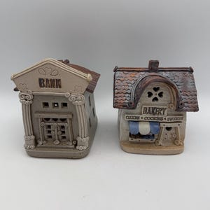 May include: Two ceramic miniature buildings: a bank and a bakery. The bank is beige with columns and the word "BANK" on the front. The bakery is tan with a blue and white striped awning and the word "BAKERY" above the entrance. The bakery also has the words "CAKES COOKIES & SWEETS".