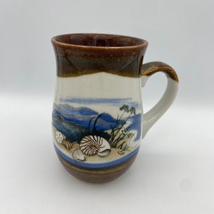 May include: A brown and white ceramic mug with a seashell design. The mug has a brown rim and handle. The design features a seashell, a starfish, and a seascape.