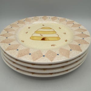 May include: A stack of four round ceramic plates featuring a central illustration of a yellow beehive surrounded by small bees. The rim displays a repeating pattern of beige diamonds accented with small blue dots on a cream-colored background.