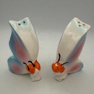 May include: A pair of ceramic butterfly-shaped salt and pepper shakers. The wings are white, with blue and pink accents. The butterfly bodies are orange and white, with black antennae. Each shaker has three holes.