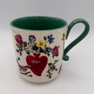 May include: A ceramic mug with a green handle and rim. The mug features a red heart-shaped vase filled with flowers and the word "LOVE". Colorful birds and floral accents decorate the off-white surface.